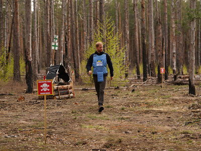 Prince Harry, wearing a HALO vest with the Ukrainian flag, walks through a wooded area featuring a "danger: mines" sign.